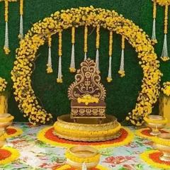 This royal Haldi seating arrangement includes an ornate chair placed inside a large urli. The backdrop is a beautiful arch made of yellow marigolds against a green wall, with hanging floral tassels adding to the charm.