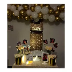 We can transform even a simple wall into a stunning feature. This date night setup includes a shimmering curtain of fairy lights, a personalized photo display table, and a beautiful gold and white balloon garland.