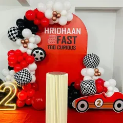 "2 Fast 2 Curious" - a high-speed race car theme for a second birthday. The decor includes a checkered flag balloon arch, a race car cutout, and a bold red backdrop.