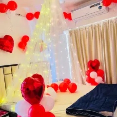 A romantic room setup with a red and white theme. A beautiful canopy made of net and fairy lights hangs over the bed, surrounded by red and white balloons and heart-shaped foil balloons, creating a dreamy ambiance.