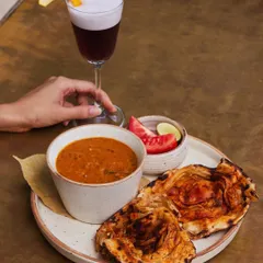 Comfort in a bowl. Our hearty dal is served with flaky parathas and a side of fresh tomato and lime, perfectly complemented by a frothy, dark cocktail.