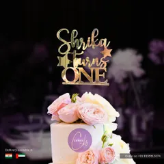 A "Shrika Turns ONE" topper in glossy gold acrylic, shown here on a simple white cake adorned with fresh pink roses. This style combines classic elegance with a personal touch.