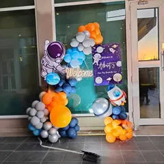 A welcome setup for a space-themed party. This arrangement includes an astronaut cutout, a personalized welcome sign, and a balloon garland in orange, blue, and grey.