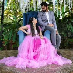 A couple enjoys a regal themed pre-wedding shoot. The client is seated in a blue throne chair, wearing our bright pink ruffled gown.