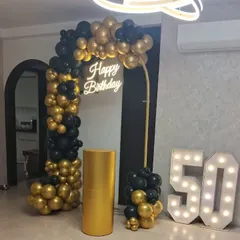 An elegant and modern 50th birthday party decoration. The setup includes a black and gold chrome balloon arch, a 'Happy Birthday' neon sign, and large light-up marquee numbers to mark the milestone.