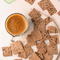 My Ragi Crackers, scattered on a marble surface next to a cup of coffee. Baked, not fried, these light and crispy crackers are packed with the goodness of ragi and are perfect for guilt-free munching.