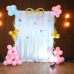 A sweet and simple backdrop for a celebration at home. This setup features a white curtain with hanging photos, framed by small balloon bunches in pink, blue, and gold.