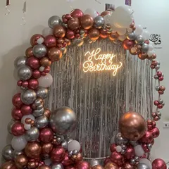 This is how we bring the party to you. We created this luxe balloon arch for a special birthday celebration at home, using a mix of metallic rose gold, silver, and white balloons, complete with a 'Happy Birthday' neon sign against a silver fringe curtain.