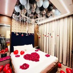 An elegant bedroom decoration featuring silver, black, and light blue balloons on the ceiling. The bed is adorned with a large heart made of red rose petals, and photos of the couple hang from the balloon strings for a personal touch.