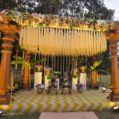 A stunning South Indian theme Haldi stage. The mandap is built with traditional wooden pillars, a canopy of jasmine flowers, and a vibrant yellow and white chevron-patterned floor.