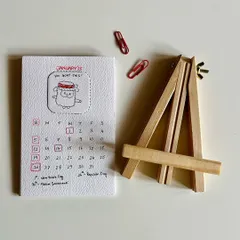 The full 2025 desk calendar set, showing the January page, the wooden easel, and the red clips used to hold the pages in place.