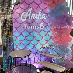 A personalized mermaid-themed backdrop for Anika's 6th birthday. The design features a shimmering scale pattern, the birthday girl's name, and a beautiful balloon garland in pastel pinks and blues.
