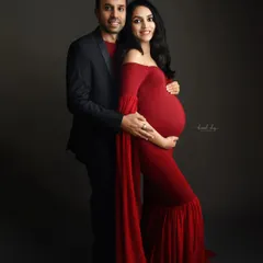 A full-length formal portrait of an expecting couple. The striking red gown with its long, flowing sleeves stands out beautifully against the moody, dark background of the studio.