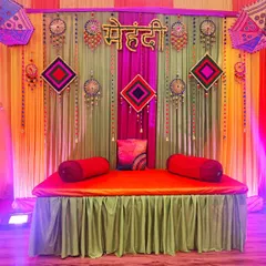 For a Mehendi night, I can create a stunning backdrop like this one. It includes a comfortable diwan for the bride, decorated with colourful drapes, traditional hangings, and Rajasthani umbrellas for beautiful photos.
