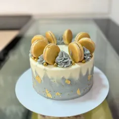 A stylish grey and white watercolor cake. This design is elegantly decorated with gold leaf, grey buttercream swirls, and a crown of golden macarons.