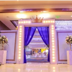 A grand entrance fit for a king. This royal-themed party welcome features a marquee-lit archway topped with a crown and draped with rich blue curtains, flanked by elegant floral arrangements.