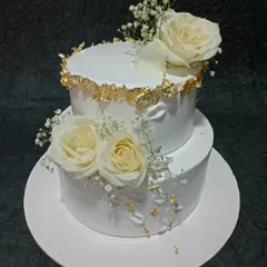An elegant two-tiered cake adorned with fresh white roses and delicate baby's breath. The torn-edge gold leaf detailing adds a touch of rustic glamour, ideal for weddings or anniversary celebrations.