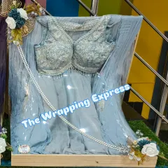 This light blue lehenga with silver embroidery is packed on a wooden base, decorated with matching blue and white flowers. It's a modern and elegant way to present a trousseau gift.