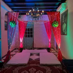 An intimate indoor mandap or ceremony setup with a low seating arrangement. The space is defined by red drapes, a beautiful chandelier, and fairy lights for a cozy and magical feel.