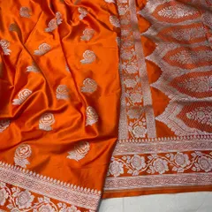 A bright orange Mashru silk saree that is perfect for festive celebrations. The silver floral weaving adds a touch of traditional elegance to the vibrant color.