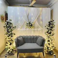 This elegant Roka setup at home features a beautiful white drape backdrop with fairy lights, flanked by two pillars of white roses. The grey velvet sofa adds a touch of modern sophistication.