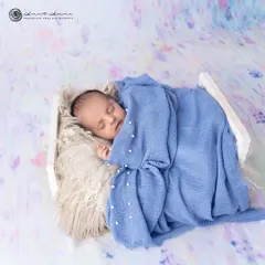 A wider shot of this peaceful newborn, showing how the dreamy, painted backdrop complements the soft blue wrap, creating a beautiful, artistic portrait.
