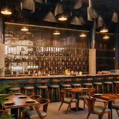 A frontal view of the main bar, the heart of the restobar. The design features a live edge wood countertop, green subway tiles, and a dramatic, back-lit bottle display. The expanded metal mesh ceiling above the bar adds another layer of industrial texture.