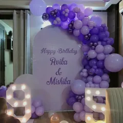 A beautiful purple-themed decoration for a joint birthday celebration. The custom arched backdrop is framed by a balloon garland in various shades of purple, complemented by large light-up numbers '9' and '5'.