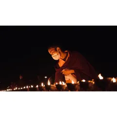 A monk wearing a mask lights a diya during a lamp offering ceremony. This close-up focuses on a single act of devotion, highlighting the personal and meditative aspects of the larger event.