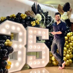 The birthday boy posing with his custom 35th birthday decor. This setup includes 4-foot tall marquee numbers surrounded by a lavish black and gold balloon arrangement with decorative dried palms.