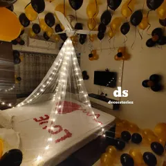 An elegant black and gold themed room decoration for a newly married couple. The setup includes a light-up canopy, 'LOVE' written in rose petals on the bed, and balloons covering the ceiling and floor.