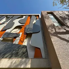 The "Framed Austerity" artwork seen in context with the building's textured facade. The organic, flowing lines of the metal art create a dynamic contrast with the rigid structure, while the tree's shadow adds another layer of natural pattern.