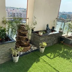 This balcony makeover features a stacked stone water fountain and a custom-built planter box with a Shiva statue, creating a spiritual corner. The use of artificial turf and potted plants adds softness and life to the compact space.