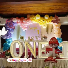 A vibrant rainbow butterfly theme for Lasya's first birthday. This stage setup includes a large 'ONE' table, mushroom props, and a colorful balloon arch.