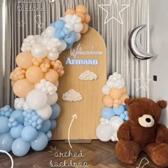 A classic "Welcome Armaan" setup featuring a teddy bear and a dreamy sky theme. The decor includes a custom arched backdrop, a beautiful organic balloon garland in peach, blue, and white, and cute cloud and moon accents.