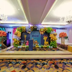 A grand jungle theme stage setup for Anant's Mundan ceremony in a banquet hall. The decor includes a large balloon arch with animal foil balloons, a personalized backdrop, and cute animal cutouts.