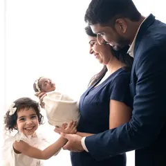 A happy family of four welcomes their newborn princess. The older sister's bright smile shows the joy of becoming a big sister in this classic, high-key portrait.