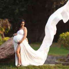 A wide shot of an expecting mother in a white gown with a long, flowing train, set against a natural, rocky landscape. This outdoor maternity photo has a beautiful, breezy feel.
