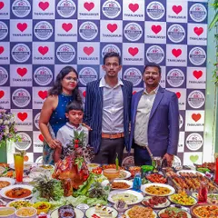 A family poses in front of our grand feast, highlighting how our catering turns any event into a memorable celebration. The table is packed with everything from biryani and curries to kebabs and desserts.