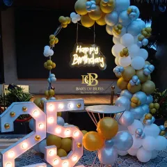 An outdoor 25th birthday celebration with a yellow, silver, and white color scheme. The decor features a balloon arch, a 'Happy Birthday' neon sign, and large marquee numbers that stand out beautifully against the evening sky.