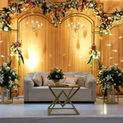 This elegant reception stage features a classic design with a gold metal framework, a wood-paneled backdrop, and abundant floral arrangements. The crystal chandeliers add a touch of timeless glamour to the setting.