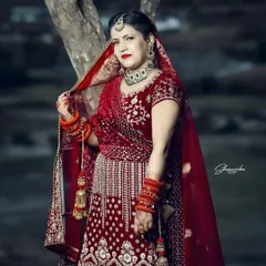 A full-length portrait of a bride in her stunning red velvet lehenga. The outdoor, natural setting allows the richness of the fabric and embroidery to truly shine.