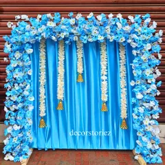 A serene blue and white themed ready-to-install backdrop for Ganpati. The design features a blue satin curtain, hanging jasmine garlands with temple bells, and a frame of artificial blue and white flowers.