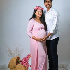 A sweet and classic studio portrait of an expecting couple. The mom-to-be wears a soft pink gown and floral crown, standing next to her partner with a vintage-style pram prop nearby.