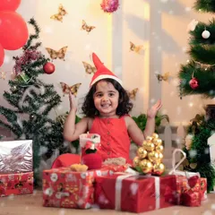A happy girl in a Santa hat sits amongst a pile of presents in a festive Christmas studio setup.