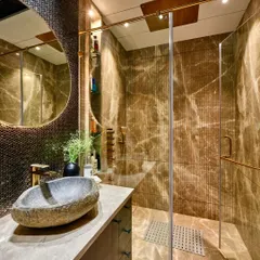 This bathroom combines rustic and modern elements. It features a natural stone vessel sink, a unique penny-tile backsplash, and fluted marble walls within the glass-enclosed shower area.