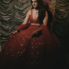 A full-length portrait of the bride seated gracefully, her magnificent red sequined lehenga spread around her. The dramatic lighting highlights the sparkle of her outfit, creating a glamorous and regal image.