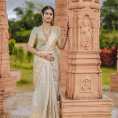 An elegant full-length portrait of a bride in a classic white and gold saree, standing beside intricately carved pillars that complement her grace.
