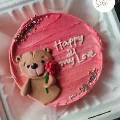 A sweet teddy bear bento cake perfect for a loved one. The fondant bear is holding a single red rose, set against a textured pink buttercream background with sparkling sprinkles.