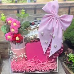 A bright and cheerful gift hamper for her, arranged in a clear acrylic box. This pink-themed set includes hair accessories, a large bow, and other cute items, perfect for a vibrant personality.